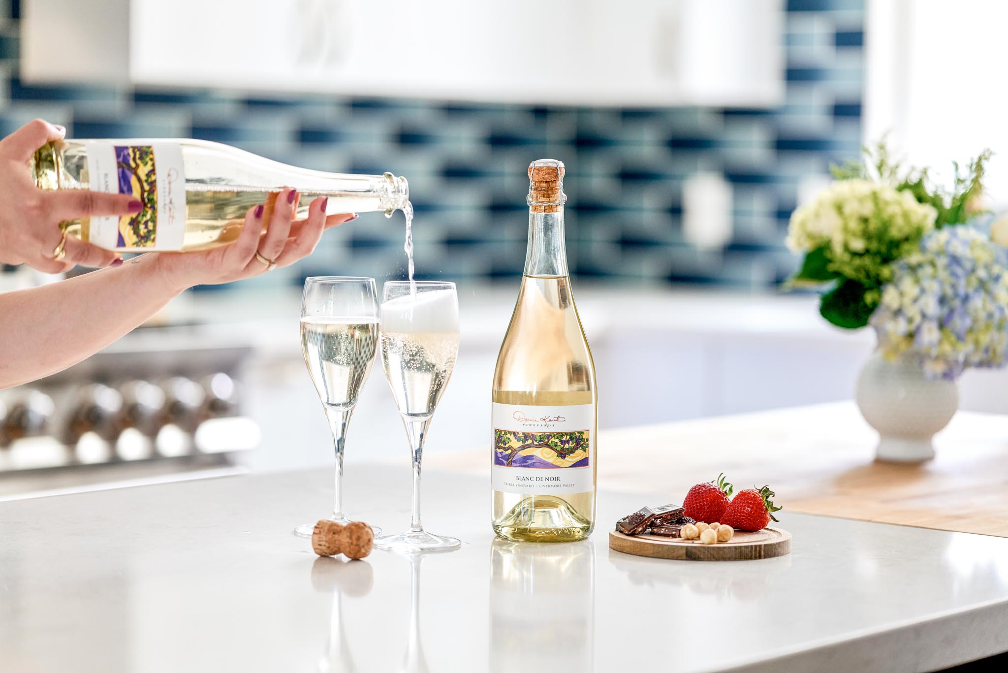 A Blanc de Noir Sparkling Wine from Darcie Kent Estate Vineyards pouring into two glasses with a small charcuterie plate on a well-lit countertop