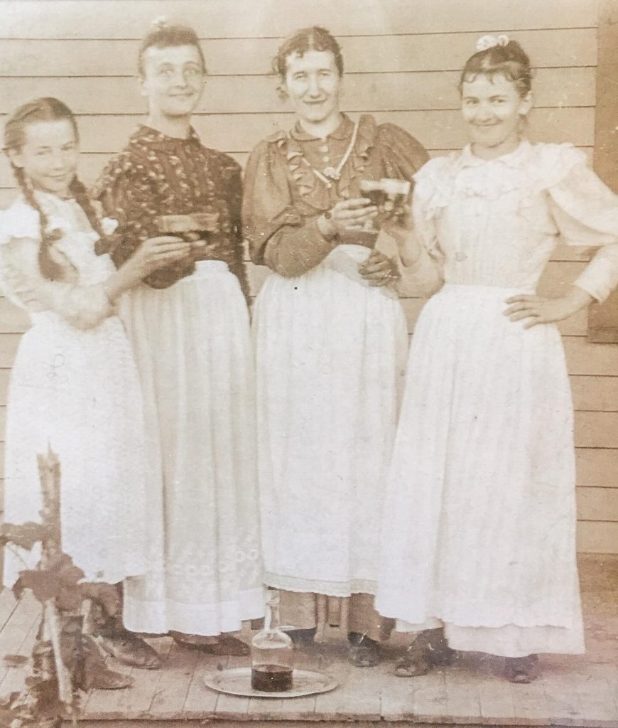 a sepia photograph of four pioneer-dressed women holding wine glasses